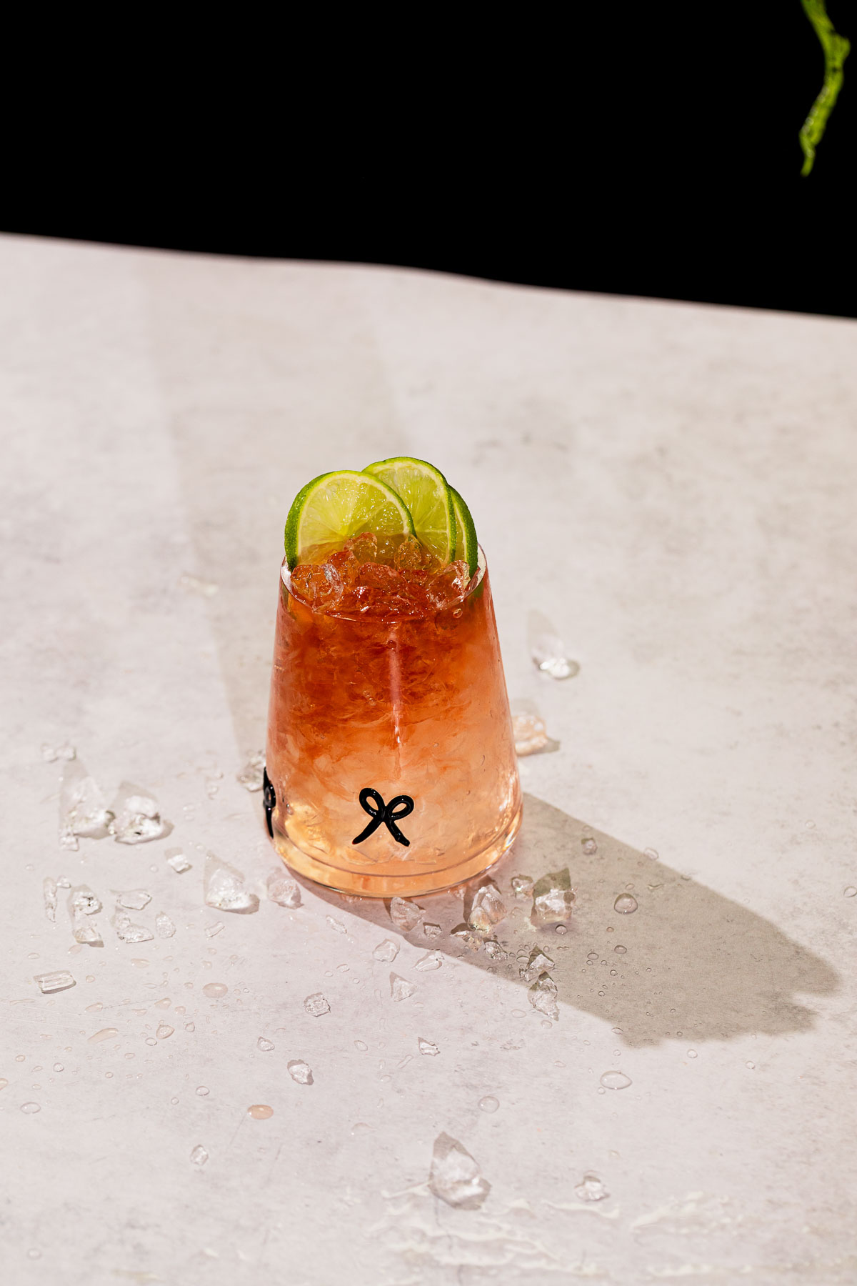 A blackberry bramble drink with crushed ice and three lime slices sits on a light surface amid scattered ice.