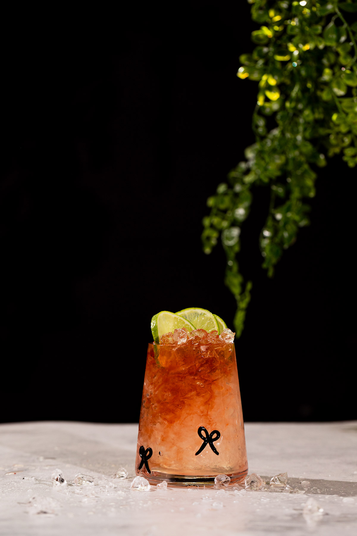 A glass with a bramble cocktail, crushed ice, and lime wedges sits on white, beneath a hanging green leafy plant.