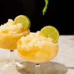 Two frozen margarita with lime wheels rest on marble beneath a hanging green plant.