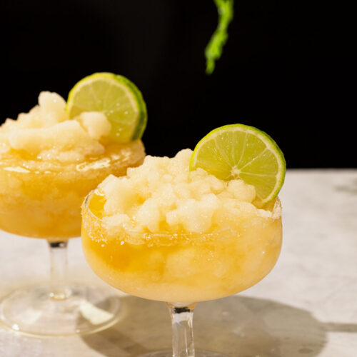 Two frozen margarita with lime wheels rest on marble beneath a hanging green plant.