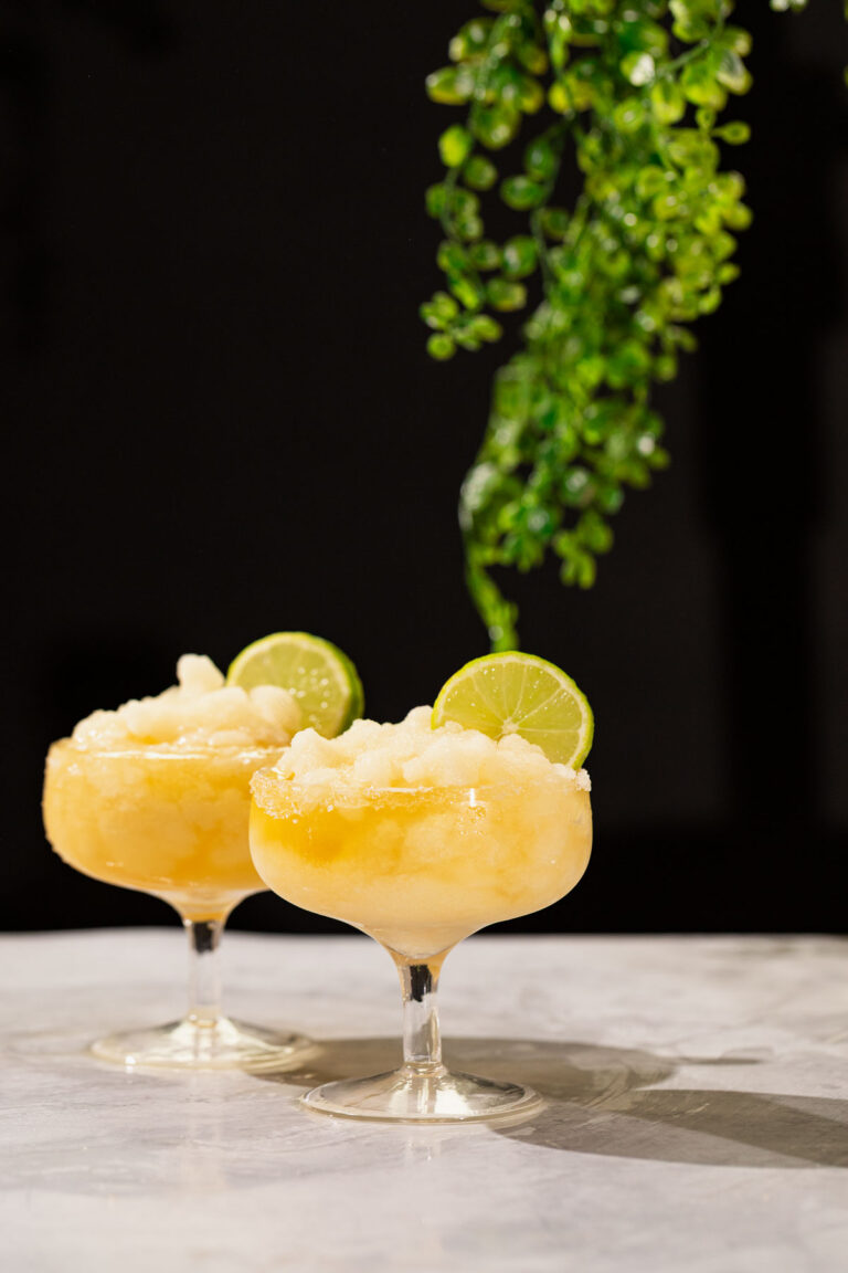 Two frozen margarita cocktails with lime slices sit on marble, with a green leafy plant hanging in the background.