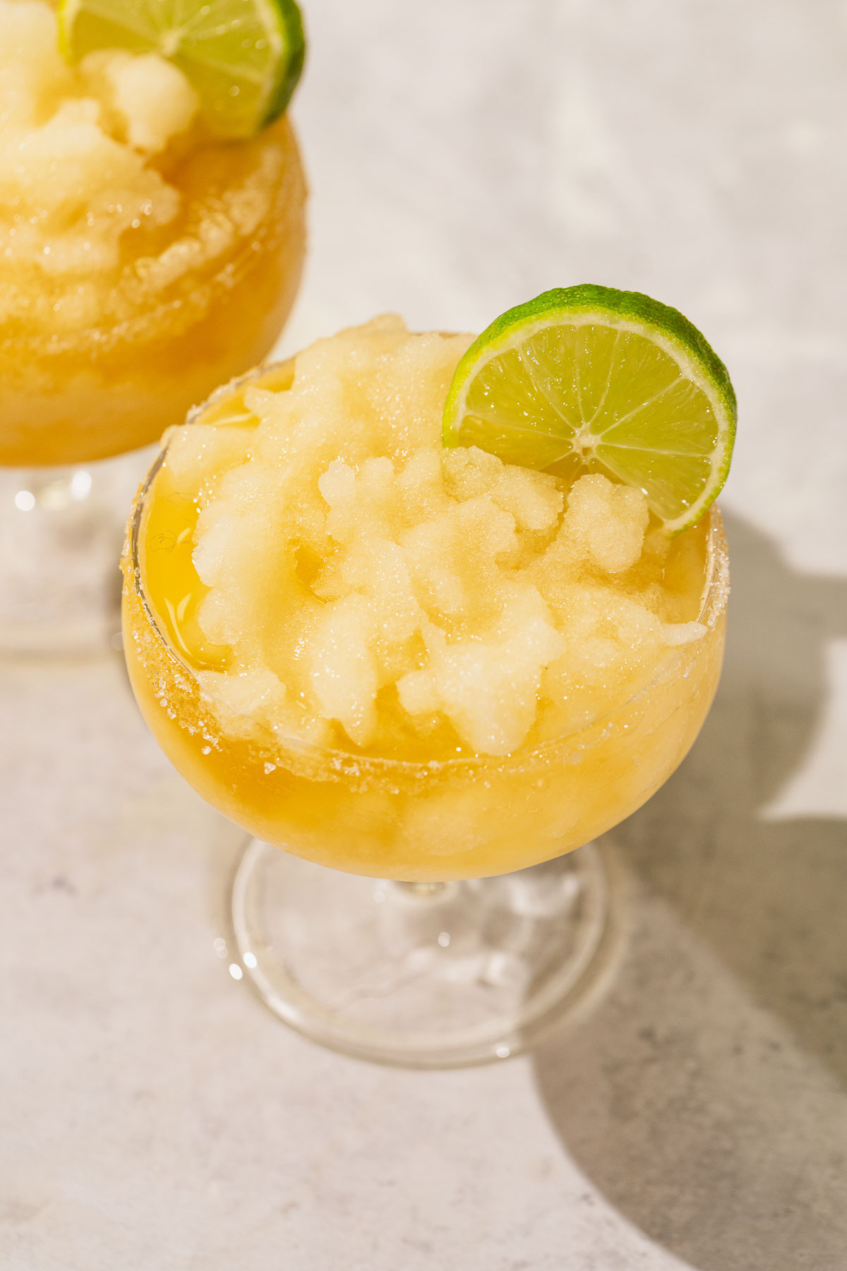 A frozen margarita cocktail in a glass with a sugared rim and lime wheel garnish rests on a light surface.