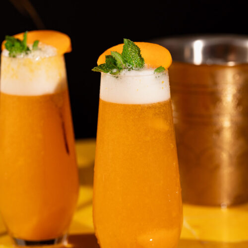 Two tall glasses of orange, frothy peach bellinis with peach slices and mint on a yellow surface, gold container in the background.