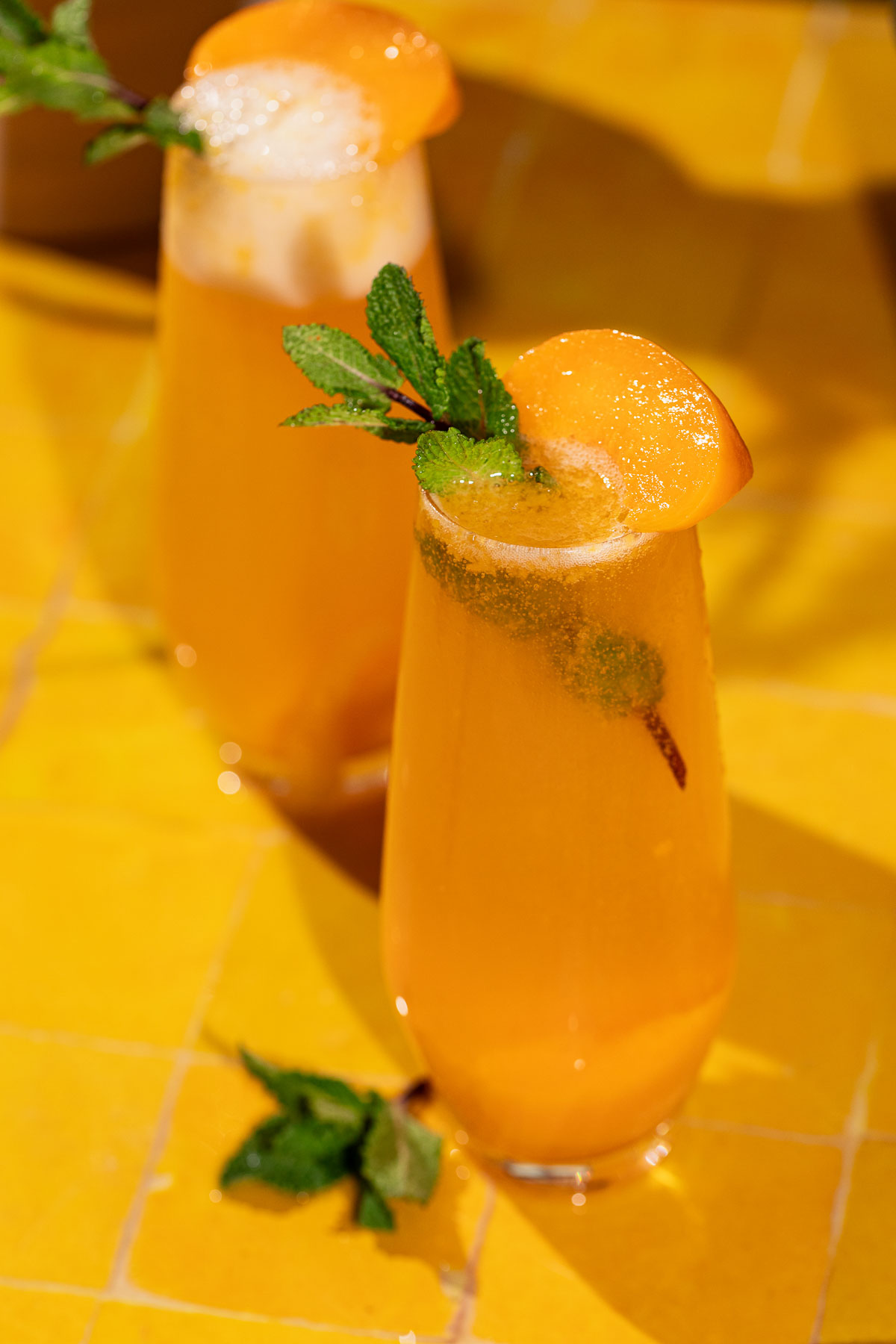 Two tall glasses of peach bellini cocktails, each topped with a peach slice and mint, sit on a yellow tiled surface.