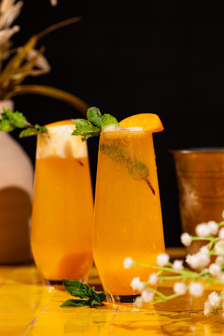 Two tall glasses of peach bellini drinks with peach and mint garnish sit on a yellow surface, decorative plants in the background.