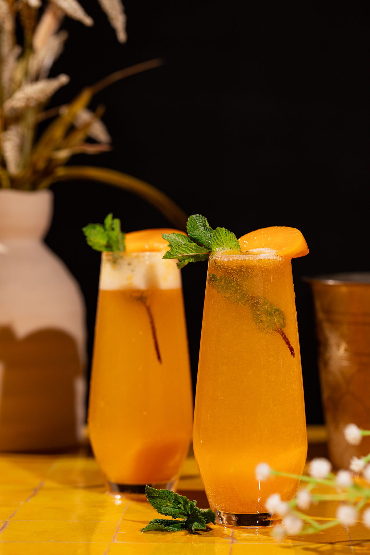 Two peach bellini cocktails with mint and peach garnishes sit on a yellow surface, with a vase and blurred background.