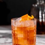 An old fashioned cocktail with ice and orange peel sits on marble, with a blurred decanter in the background.