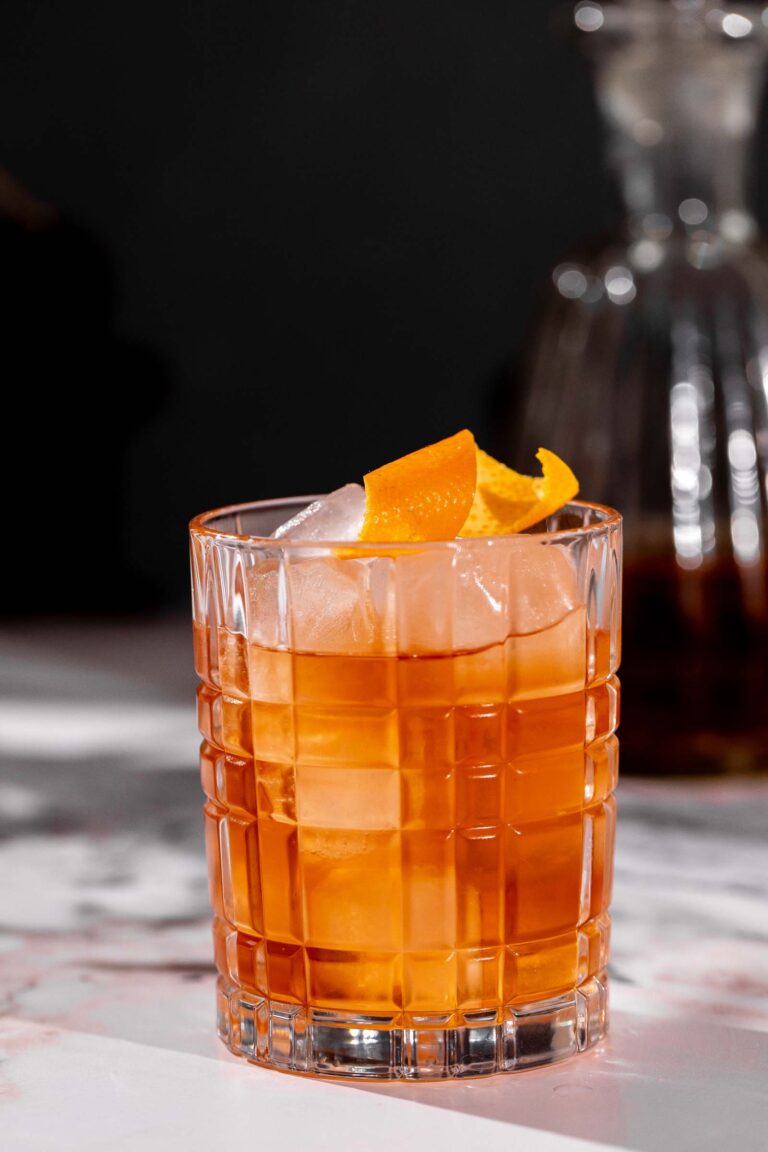 An old fashioned cocktail with ice and orange peel sits on marble, with a blurred decanter in the background.