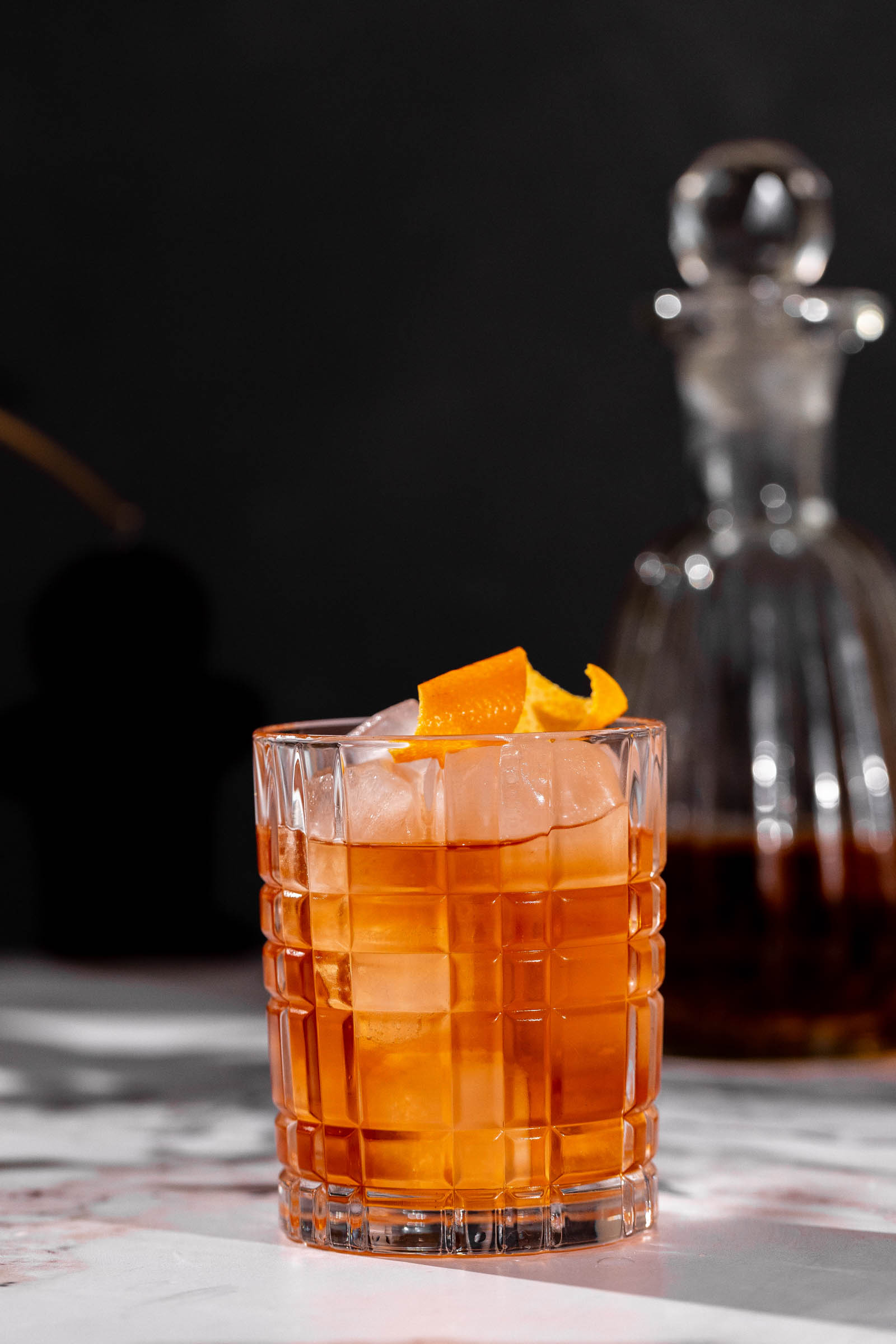 Old fashioned cocktail with ice and an orange twist sits on marble, a glass decanter visible in the background.