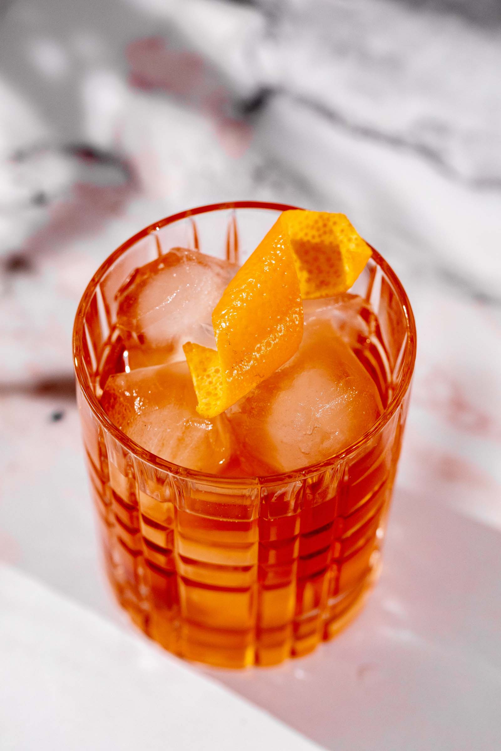 A glass of an old fashioned cocktail with ice and an orange peel garnish sits on a white surface.