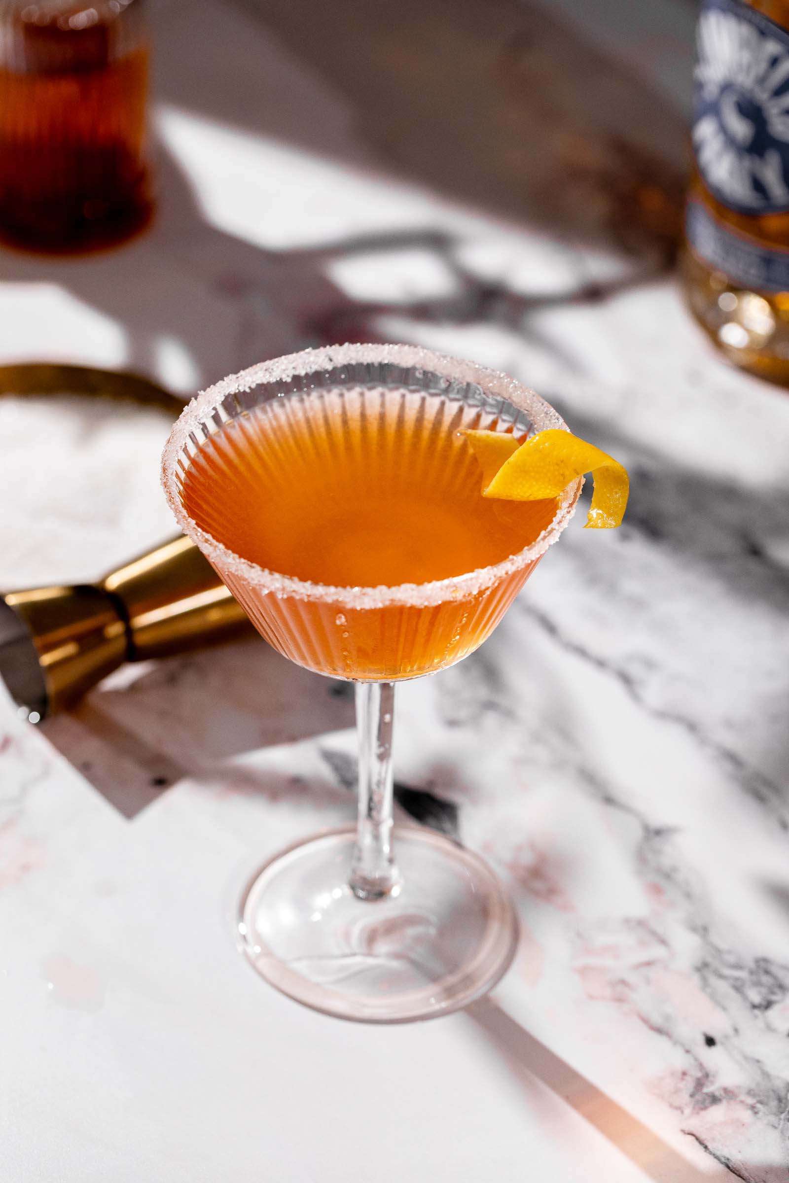A brandy crusta with a sugared rim and lemon twist sits on marble near a metal jigger, with a bottle in the background.