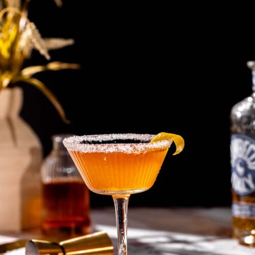 A brandy crusta with a sugared rim and lemon twist sits on marble, with a bottle, shaker, and dried wheat vase in the background.