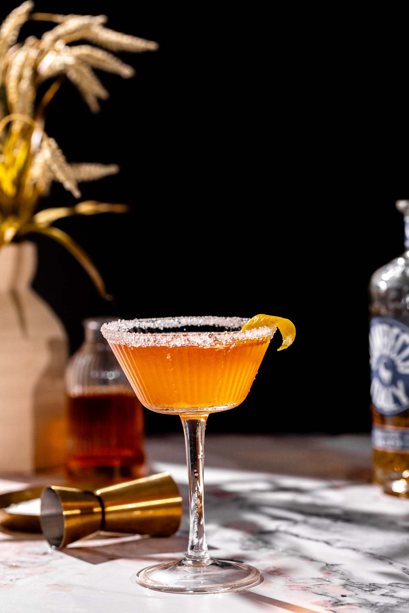 A brandy crusta with a sugared rim and lemon twist sits on marble, with a bottle, shaker, and dried wheat vase in the background.