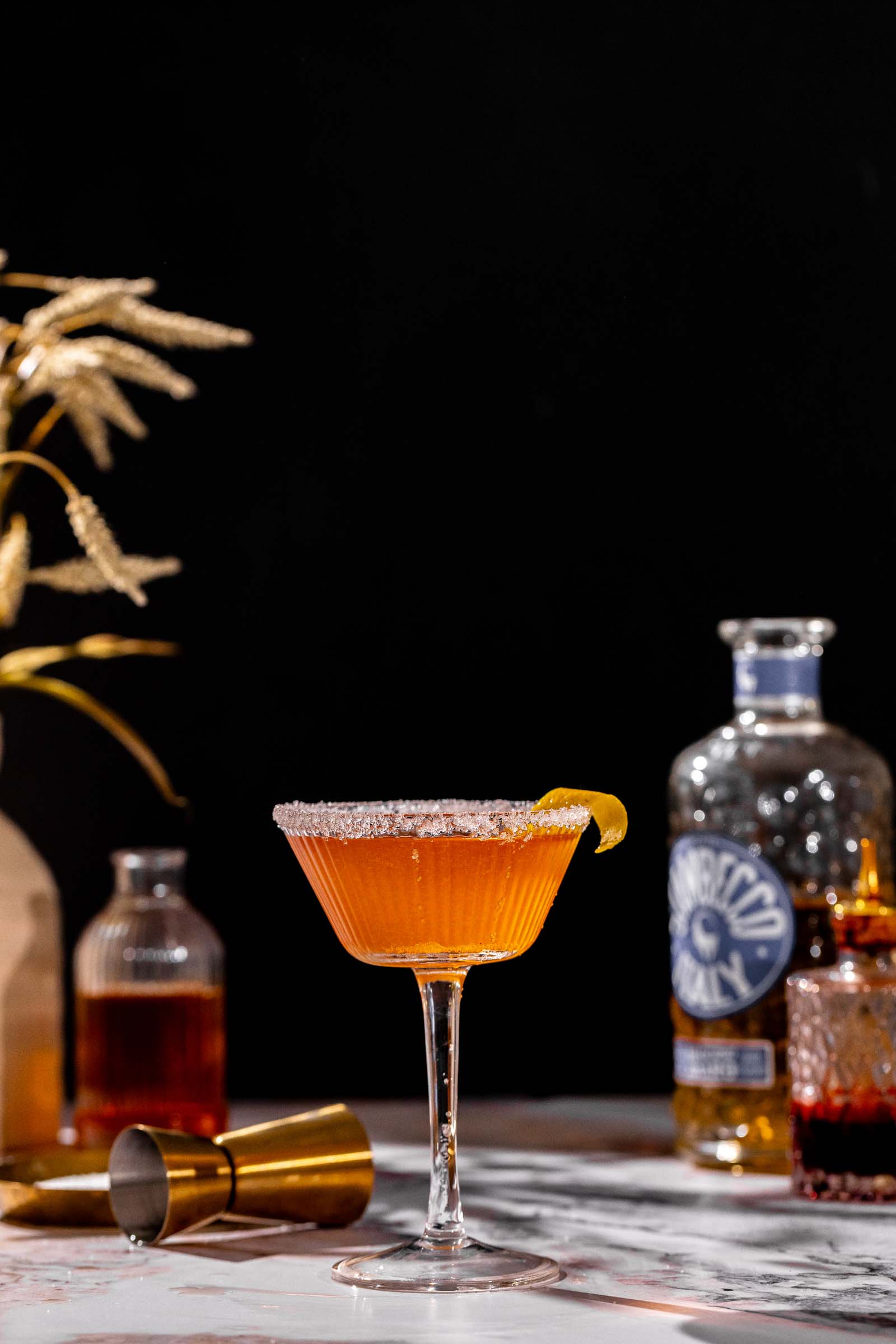 Orange-hued brandy crusta with a sugared rim and lemon twist on marble, with bottles, jigger, and dried plants in the background.