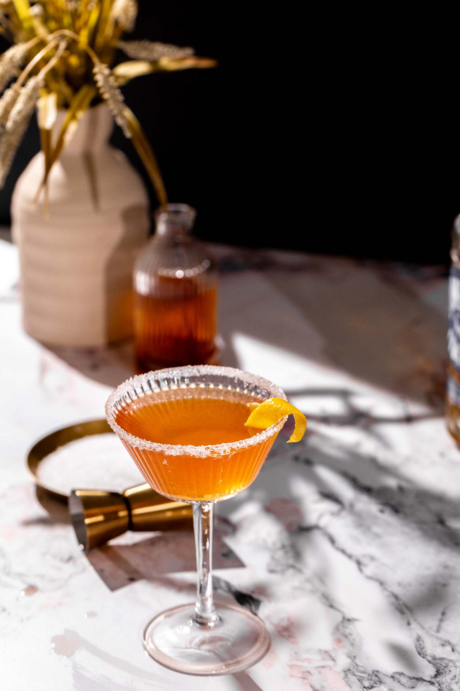 A sugared-rim brandy crusta cocktail with a lemon twist in a coupe glass sits on marble, with bottles and a vase behind it.
