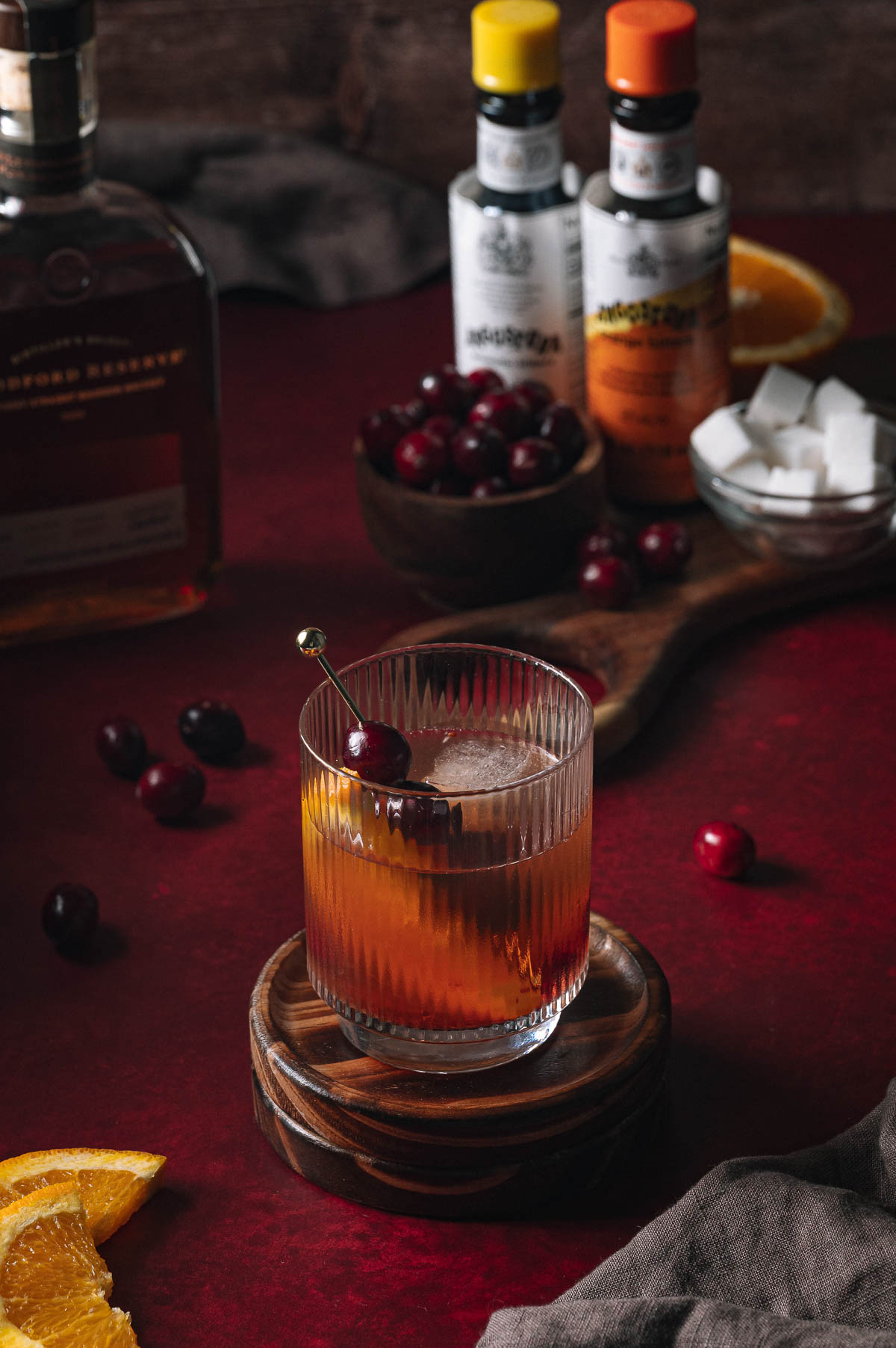 A whiskey cocktail with cherry garnish sits on a coaster amid cranberries, orange slices, sugar cubes, bitters, and a whiskey bottle.