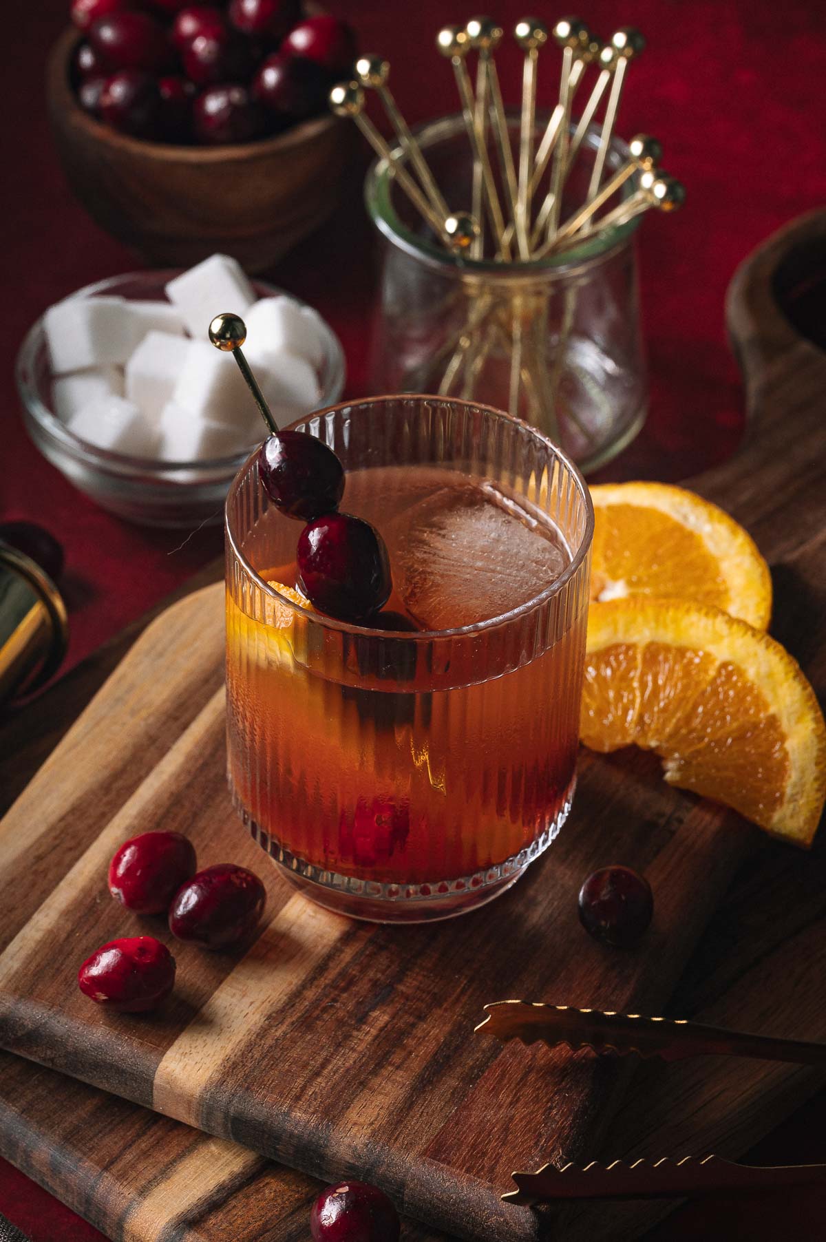 A Cranberry Old Fashioned with cherries and an orange slice sits on a wooden board surrounded by sugar cubes, fruit, and bar tools.