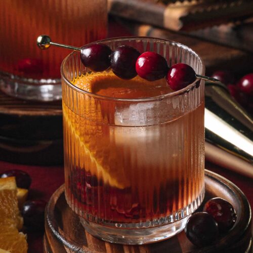 A Cranberry Old Fashioned in a glass with an orange slice and cranberries on a skewer sits on a wooden coaster beside extra fruit.
