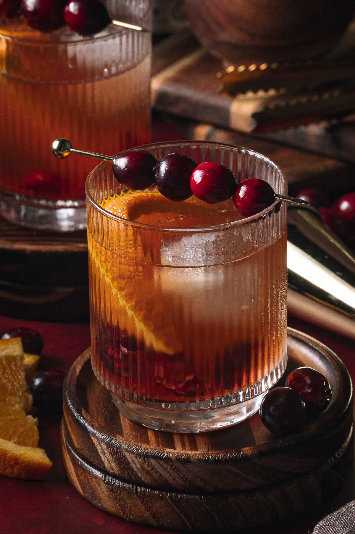 A Cranberry Old Fashioned in a glass with an orange slice and cranberries on a skewer sits on a wooden coaster beside extra fruit.