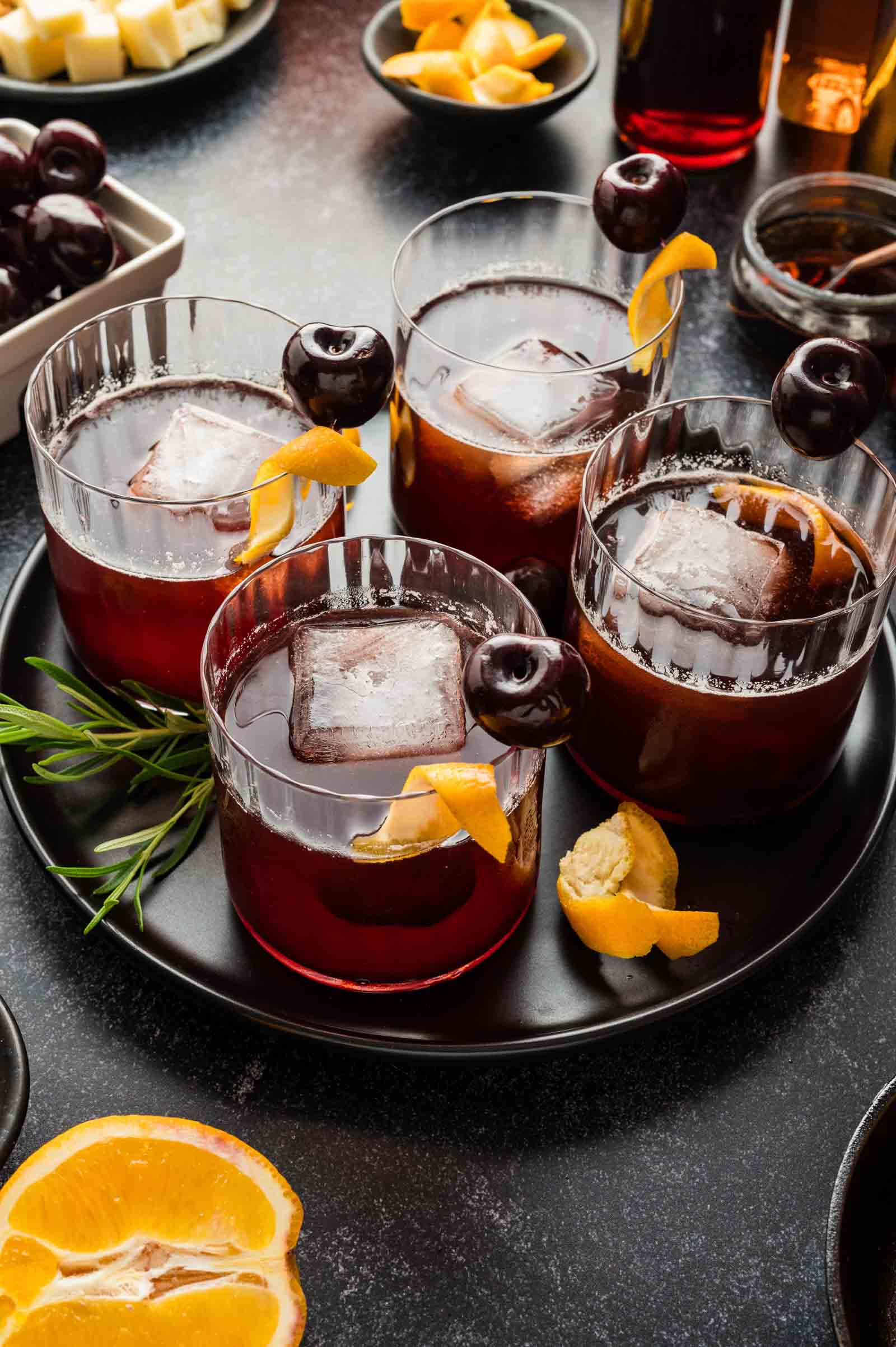 Four dark cocktails with ice, lemon peel, and cherries sit on a black plate with rosemary; fruit and snacks are in the background.