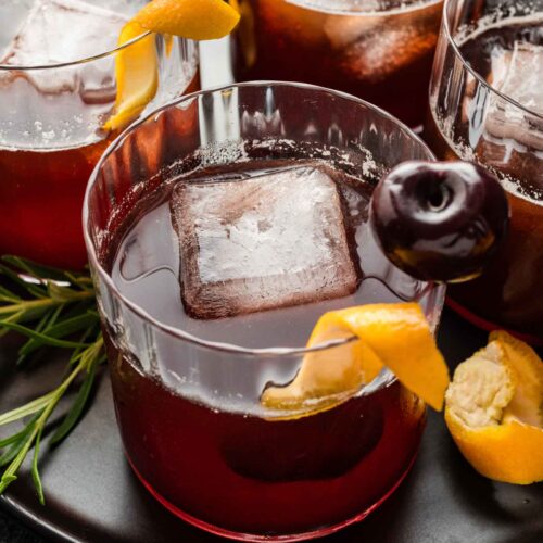 Three cocktails in glass tumblers with large ice cubes, orange peels, and dark cherries rest on a black plate.