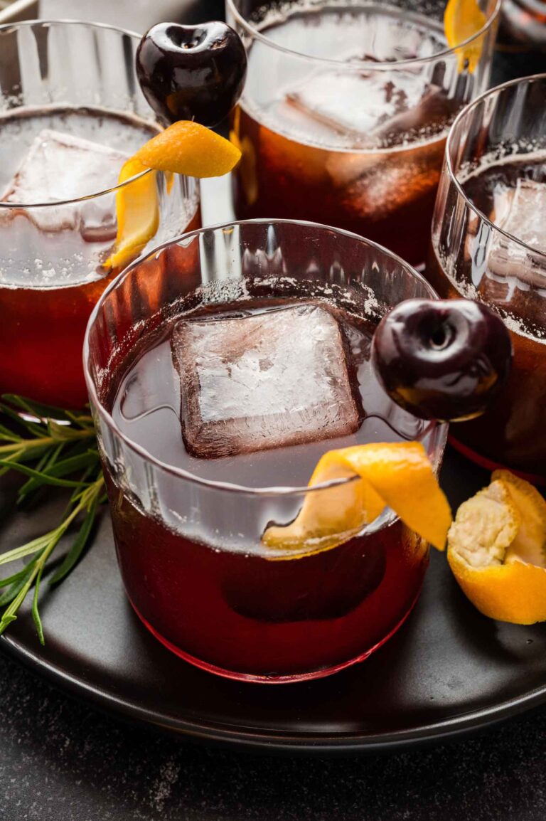 Three cocktails in glass tumblers with large ice cubes, orange peels, and dark cherries rest on a black plate.