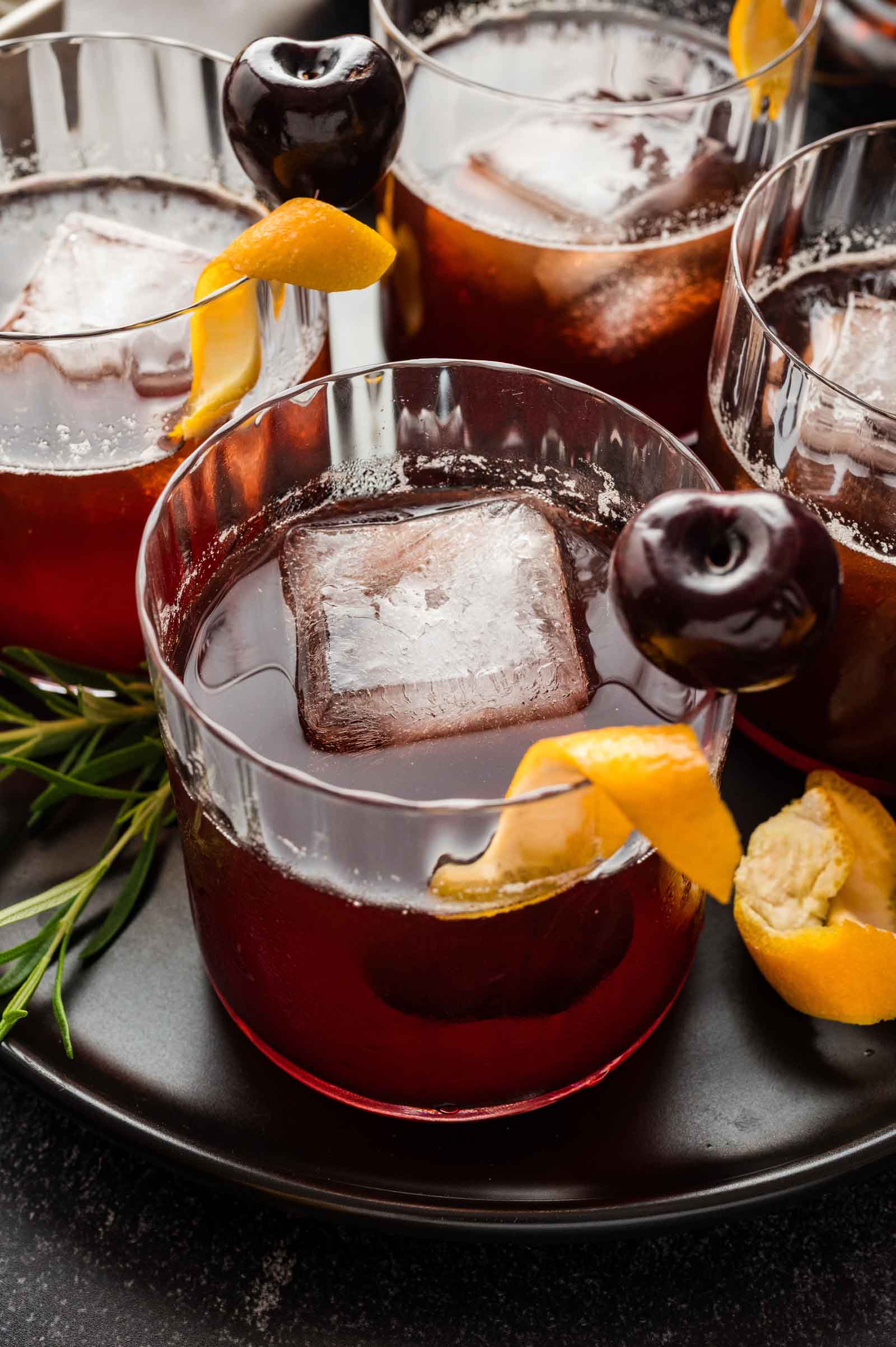 Three cocktails in glass tumblers with large ice cubes, orange peels, and dark cherries rest on a black plate.