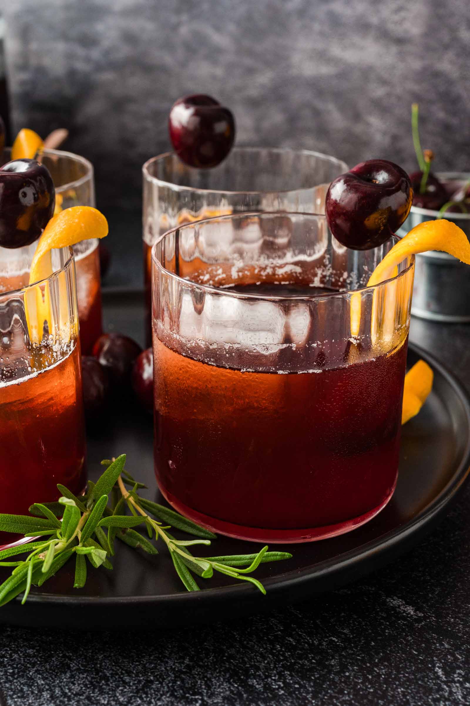 Three dark red cocktails in glasses on a black tray, topped with orange peel and cherries, with rosemary sprigs beside them.