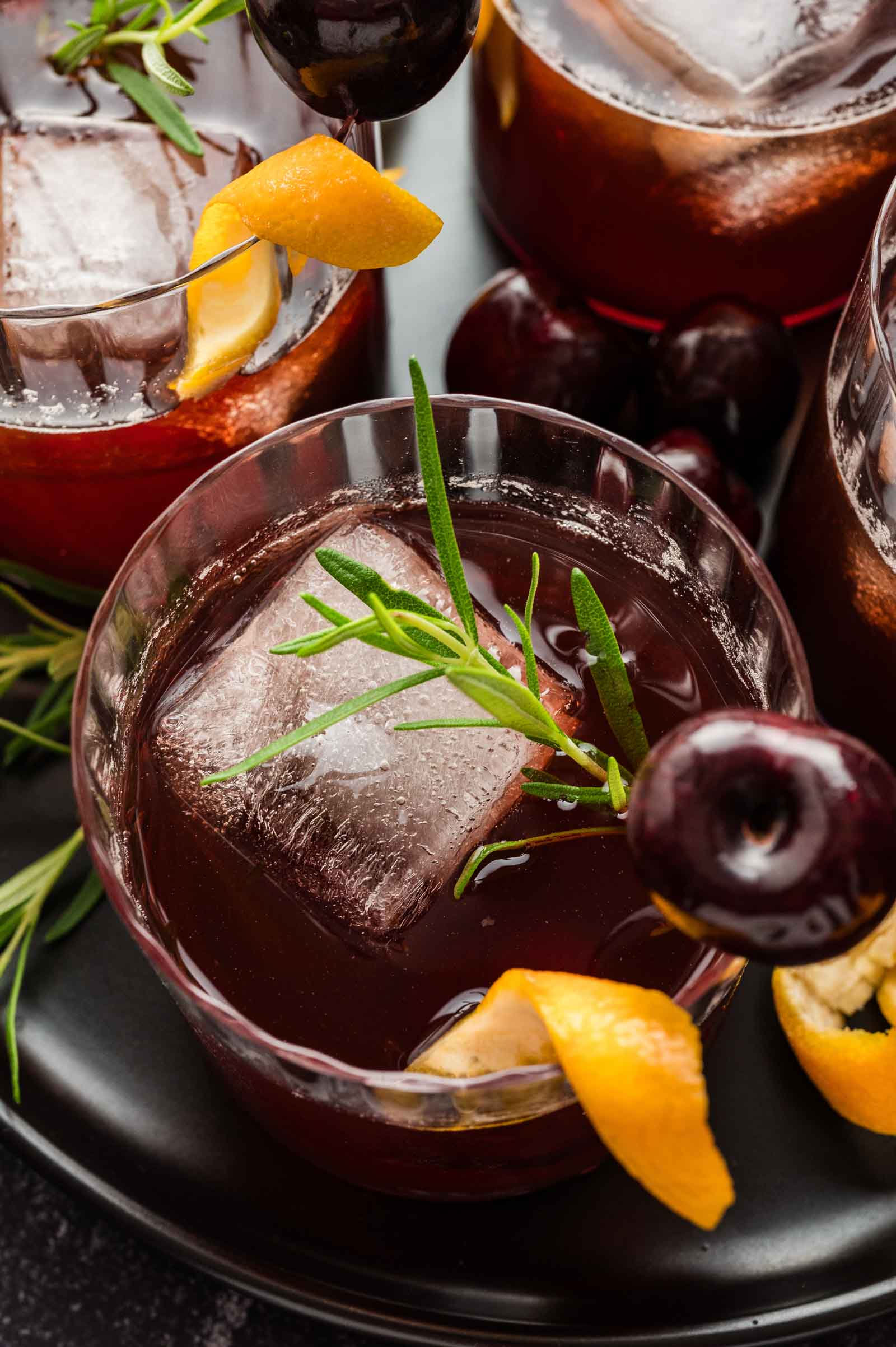 A dark cocktail with a large ice cube, cherry, orange peel, and rosemary garnish; two similar drinks appear in the background.