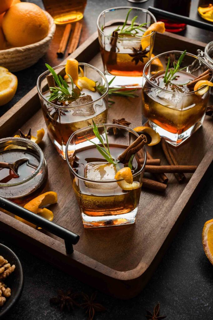Four cocktails in square glasses with ice, orange peel, rosemary, and cinnamon sticks on a tray with spices and orange slices.