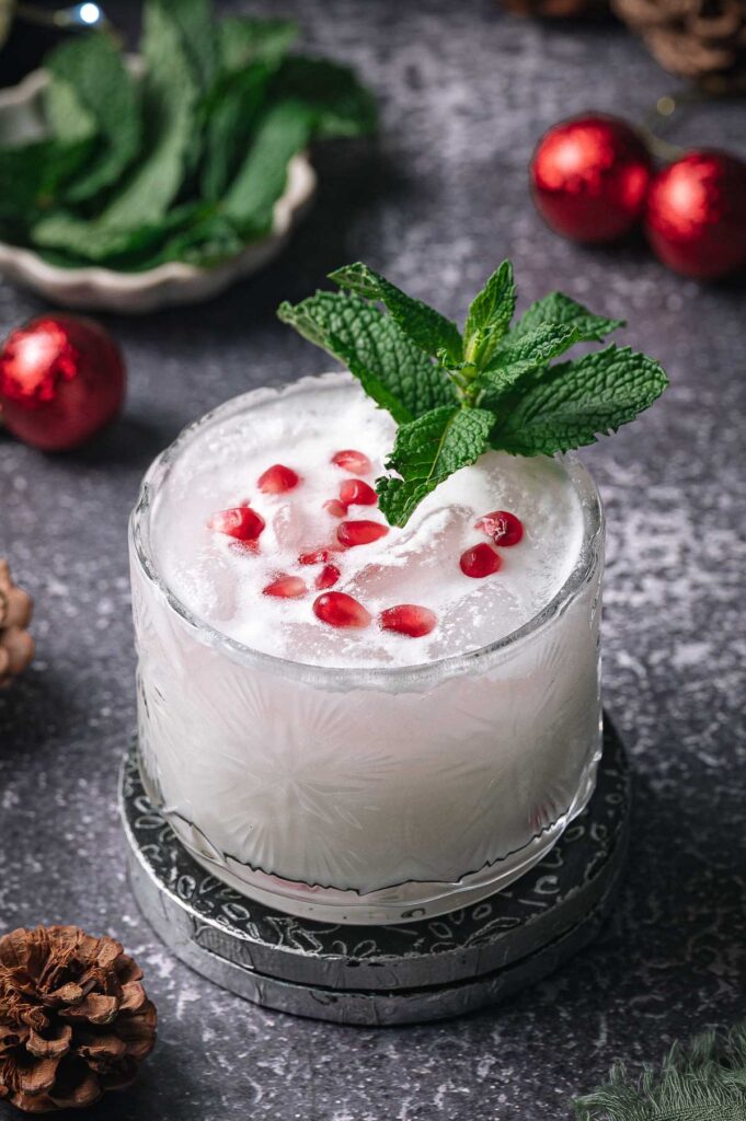 A white christmas mojito in a glass with pomegranate seeds and mint, on a coaster amid pinecones, mint leaves, and red ornaments.