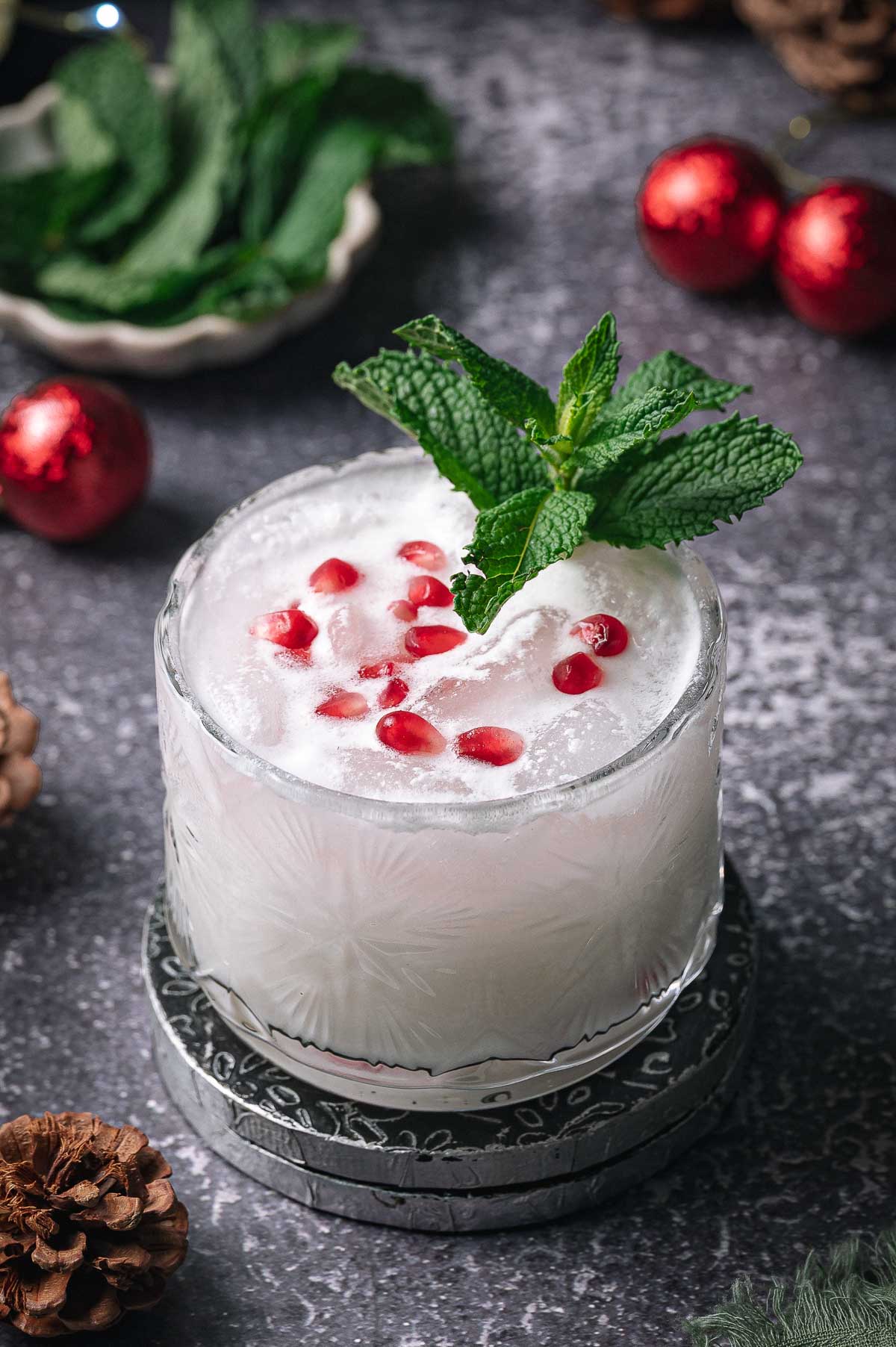 A white christmas mojito in a glass with pomegranate seeds and mint, on a coaster amid pinecones, mint leaves, and red ornaments.
