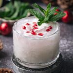 A clear glass of frothy christmas mojito cocktail with pomegranate seeds and mint sits on a coaster beside pinecones and red ornaments.