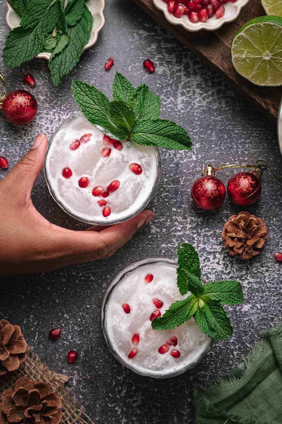 A hand holds a white christmas mojito drink with mint and pomegranate; another glass, pine cones, red ornaments, and lime are nearby.