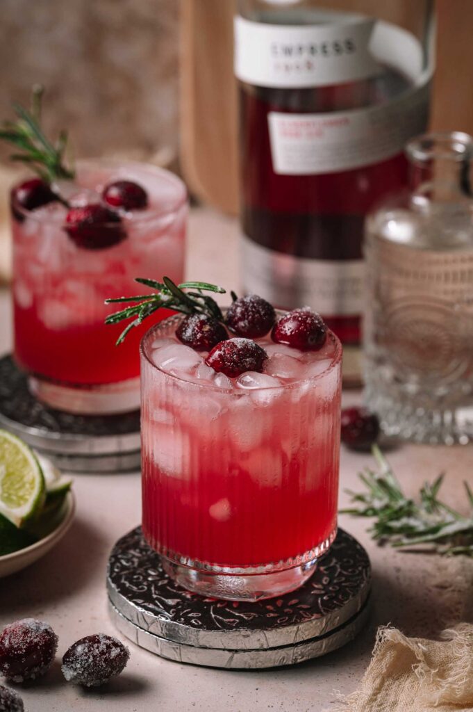 Two cranberry gin smash cocktails with ice, sugared cranberries, and rosemary on coasters, with a bottle and lime slices behind them.