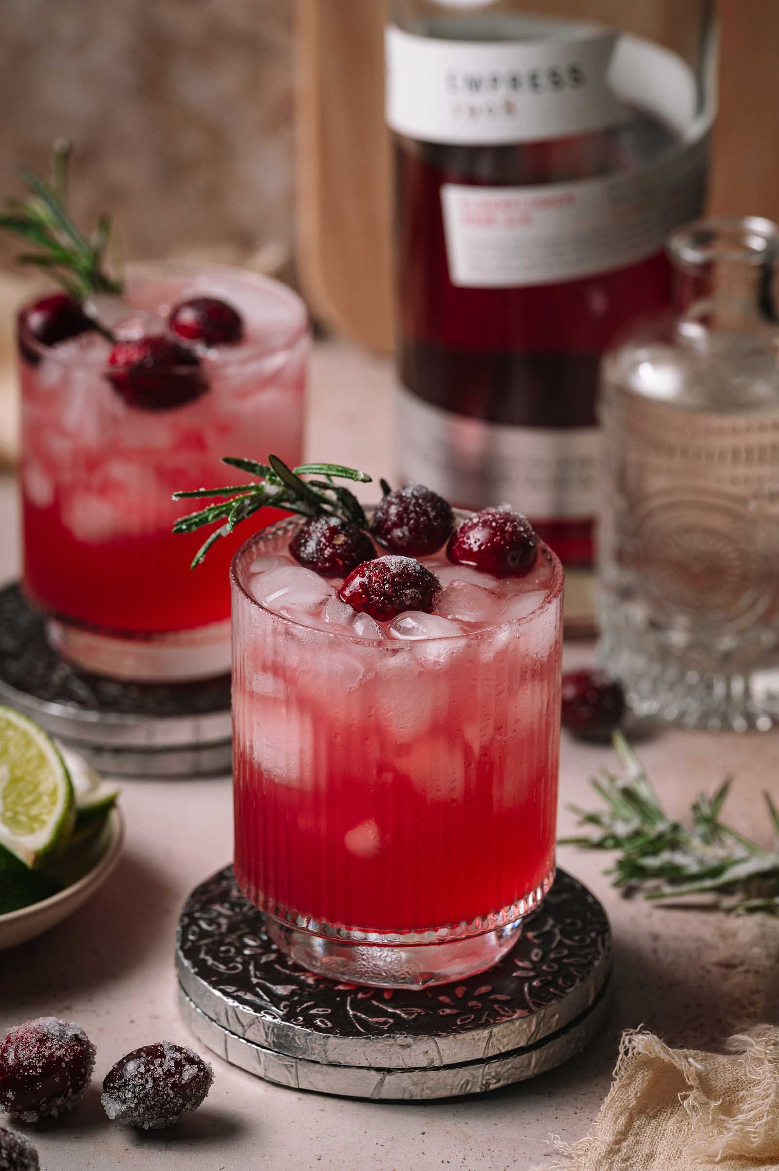 Two cranberry gin smash cocktails with ice, sugared cranberries, and rosemary on coasters, with a bottle and lime slices behind them.