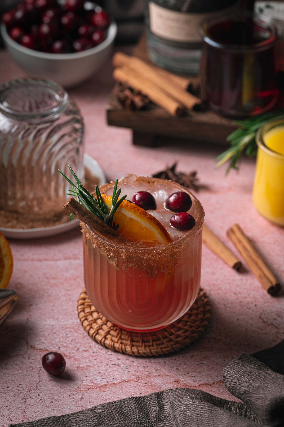 A cranberry orange margarita garnished with cranberries, orange, and rosemary on a woven coaster, surrounded by cinnamon sticks and a glass of juice.