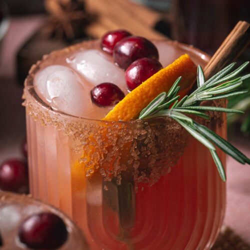 Iced pink cranberry orange margarita cocktail in a glass, topped with cranberries, orange slice, rosemary, cinnamon stick, and a brown sugar rim.