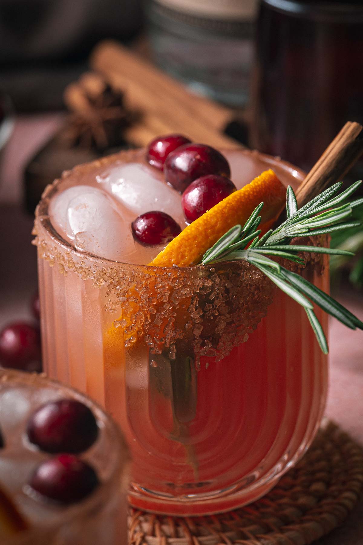 Iced pink cranberry orange margarita cocktail in a glass, topped with cranberries, orange slice, rosemary, cinnamon stick, and a brown sugar rim.