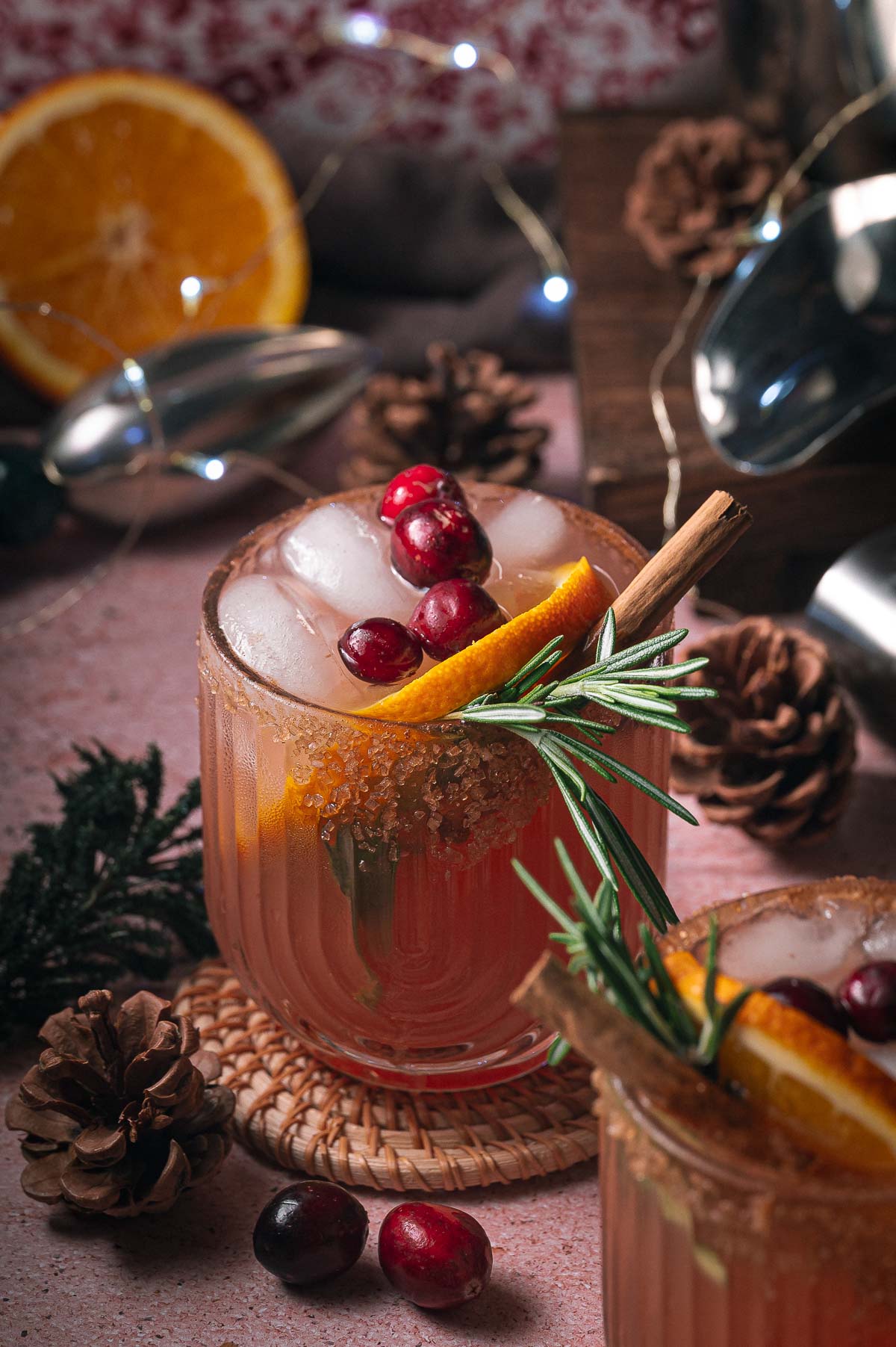 Iced cranberry orange margarita in a glass with cranberries, orange, rosemary, and cinnamon, amid pinecones and festive decorations.