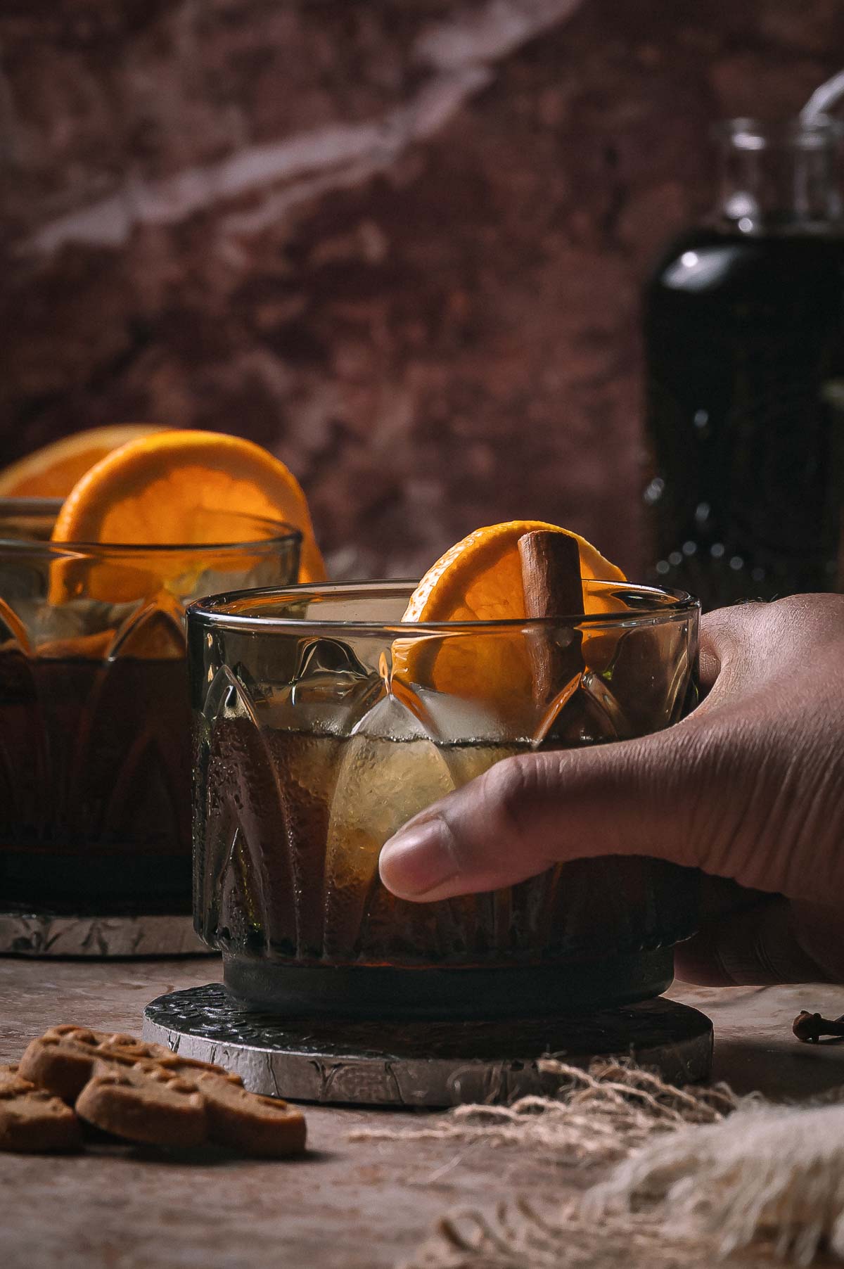 A hand holds a gingerbread old fashioned with orange slices and cinnamon on a coaster, next to another drink and cookies.