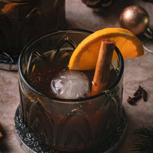 A gingerbread old fashioned drink with orange and cinnamon garnish sits on a coaster, with pinecones and holiday decor in the background.