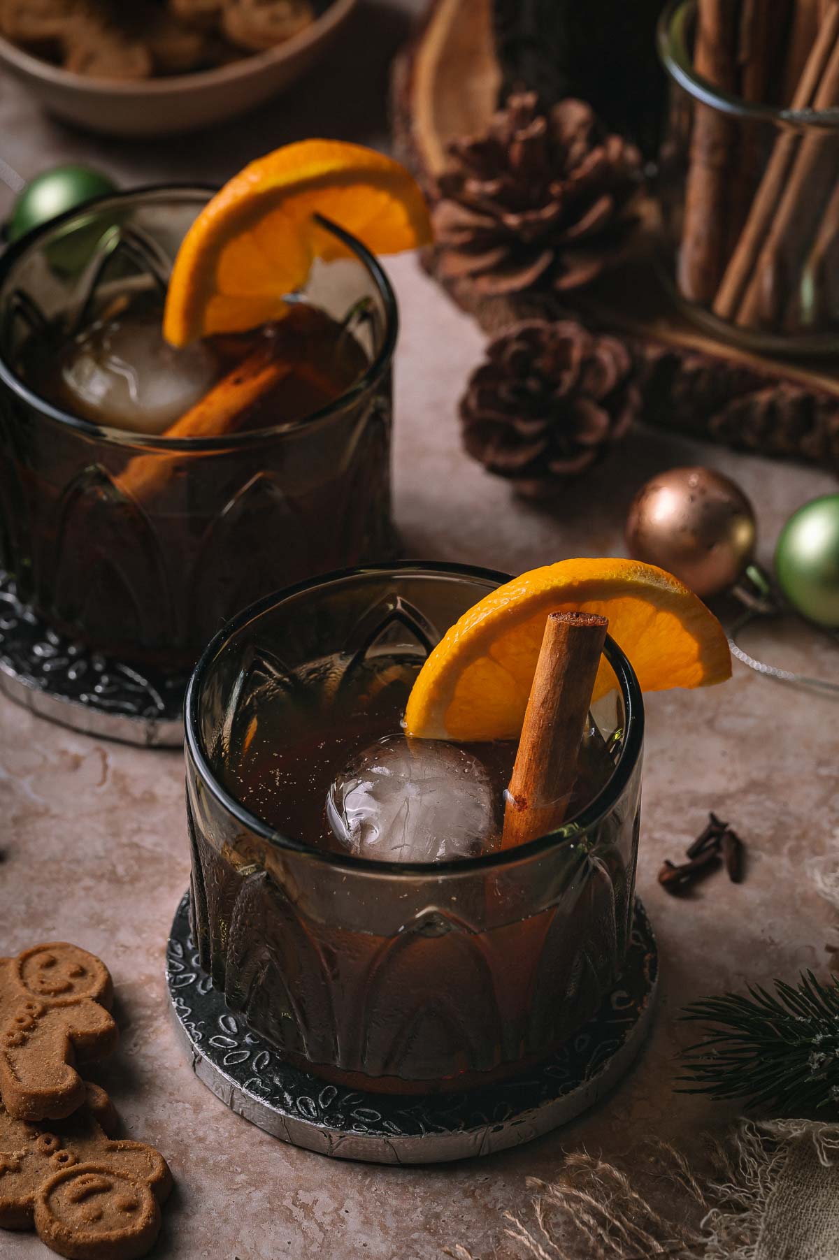 Two gingerbread old fashioneds with orange slices and cinnamon sticks sit on coasters among holiday decor and gingerbread cookies.