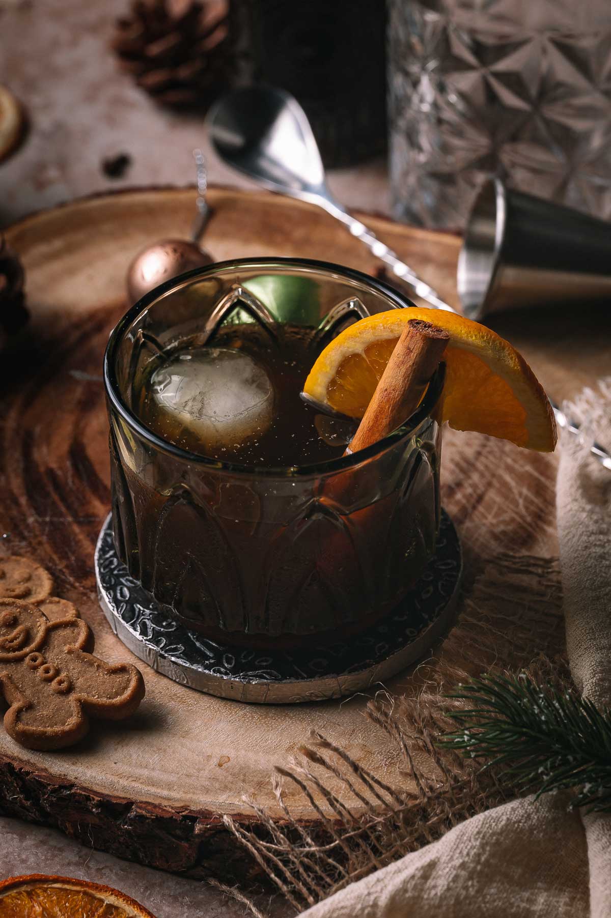 A gingerbread old fashioned cocktail with ice, orange slice, and cinnamon stick sits on wood beside a gingerbread cookie and bar tools.
