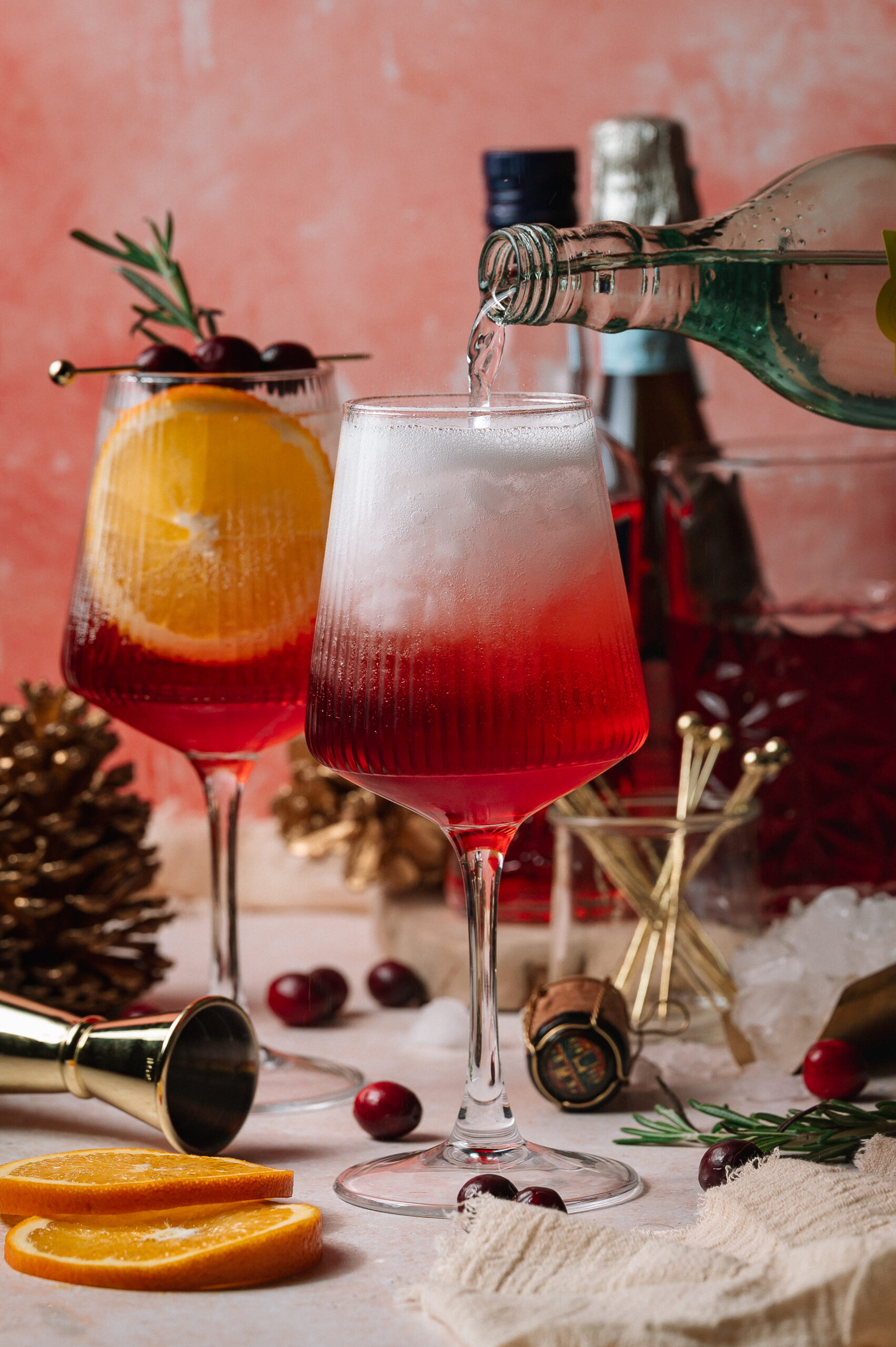 Sparkling water is poured into a red and orange Christmas aperol spritz in a stemmed glass, with another cocktail behind it garnished with orange and rosemary.