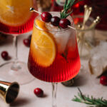 A red Christmas aperol spritz cocktail with ice, orange slice, cranberries, and rosemary sits on a table with scattered cranberries and bar tools.