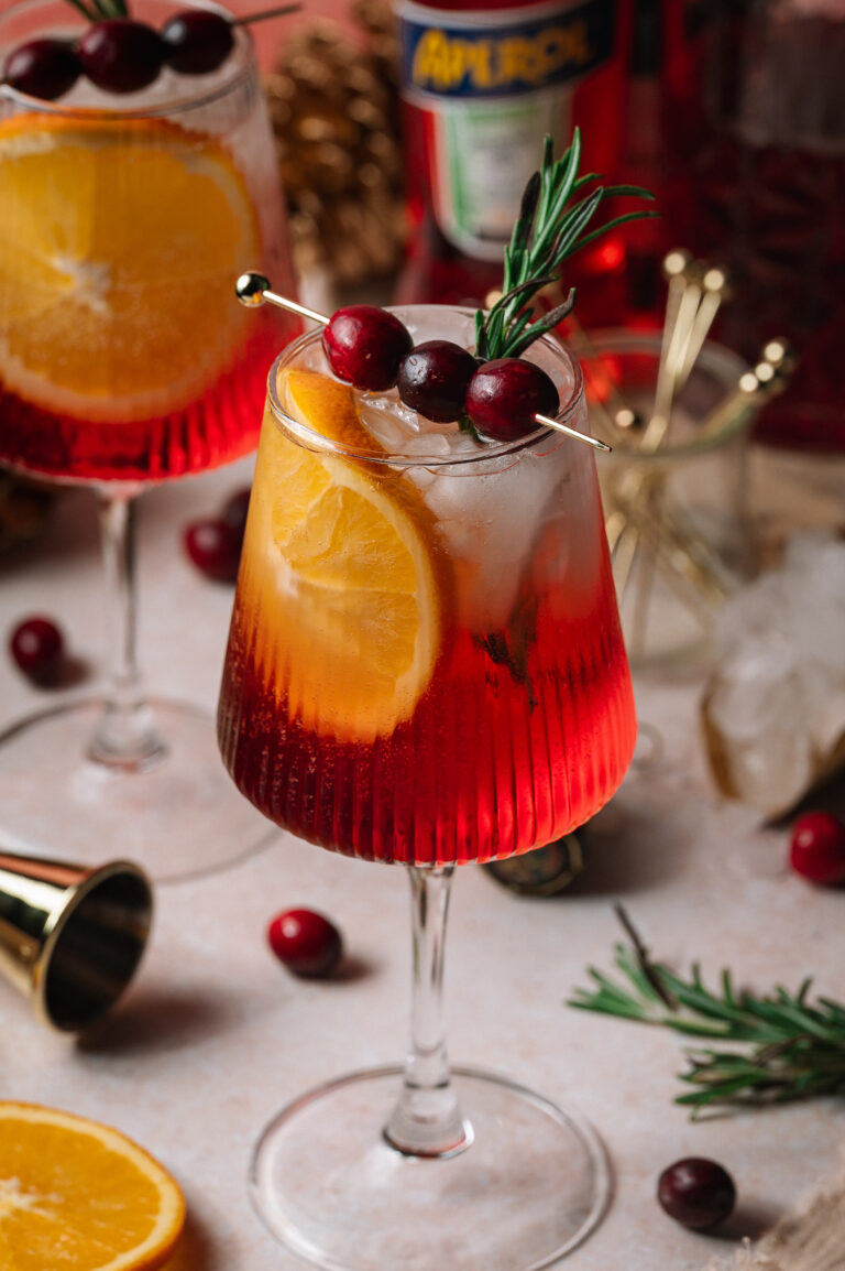 A red Christmas aperol spritz cocktail with ice, orange slice, cranberries, and rosemary sits on a table with scattered cranberries and bar tools.
