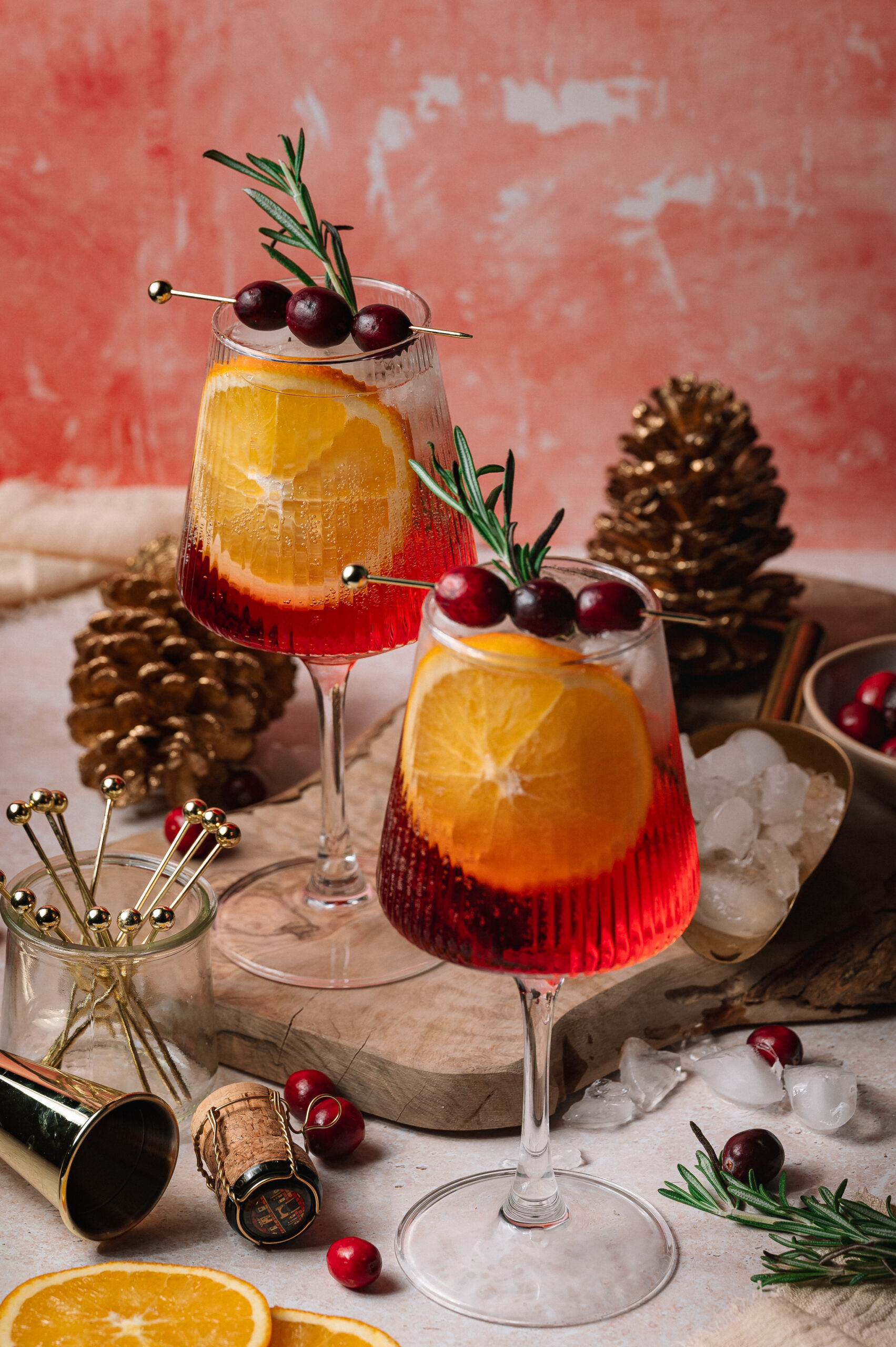 Two Christmas aperol spritz cocktails with orange slices, cranberries, and rosemary sit on ice beside gold stirrers, pinecones, and more orange slices.