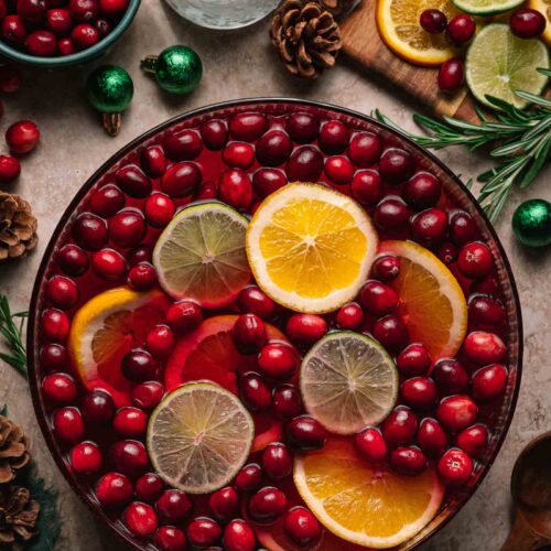 A glass bowl of festive jingle juice punch with cranberries, citrus slices, pinecones, rosemary, and two glasses of ice nearby.