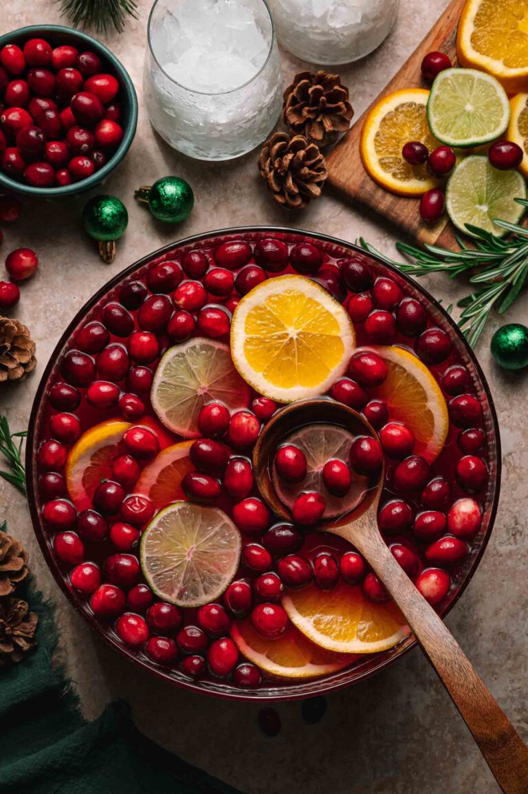 A bowl of jingle juice punch with cranberries, citrus slices, and a wooden ladle sits among garnishes and ice-filled glasses.